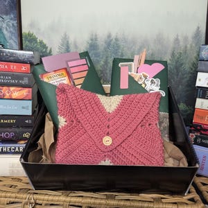 Puede incluir: Una bolsa de estilo sobre de crochet rosa y blanco con cierre de botón se encuentra en una cesta de regalo negra con una pila de libros a cada lado. Los libros tienen títulos como "The Spellshop" y "Nevernight".