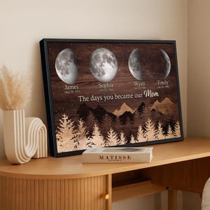 Peut inclure: Impression encadrée avec des phases de lune et des noms avec des dates en dessous. Le texte dit "The days you became our Mom". L'œuvre a un fond en grain de bois foncé avec une forêt et un design de montagne dorés.