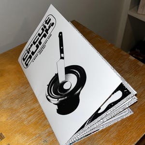 May include: A black and white magazine cover with the title "Circuit Blips" and the text "A Music Technology Zine Issue #001". The cover features a black and white illustration of a speaker with a knife sticking into it.