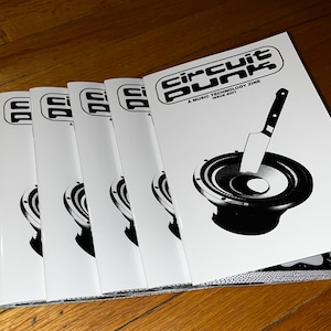 Pode incluir: Uma pilha de cinco fanzines em preto e branco intitulada "Circuit Punk: A Music Technology Zine" com o número da edição "Issue #001". A capa apresenta uma ilustração em preto e branco de um alto-falante em que um faca está enfiada.