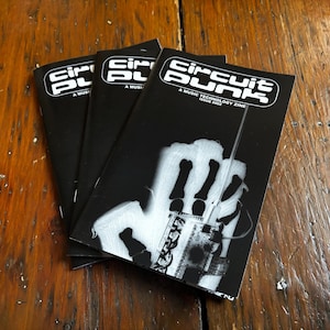 May include: Three black Circuit Punk zines are stacked on a wooden surface. The cover art features a white graphic of a hand holding a circuit board. The zines have the text "Circuit Punk" and "A Music Technology Zine" printed on the cover.