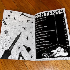 May include: An open book with a black and white cover. The book's title is "CONTENTS" and lists various topics related to guitar effects and DIY electronics. The left page features a black and white image of tools and electronic components on a dotted background.