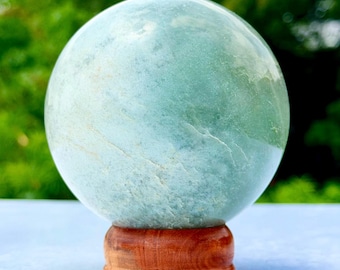 May include: A smooth, spherical stone with a pale mint green color and subtle white veining throughout. The sphere is polished to a soft sheen and rests securely on a circular, reddish-brown wooden stand with a visible natural wood grain pattern.