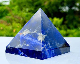 May include: A polished sodalite stone carved into a pyramid shape. The object features a deep royal blue colour with prominent white and grey mineral veining throughout its smooth, faceted surface, creating a distinct natural pattern.