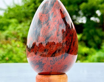 Puede incluir: Una escultura de piedra pulida en forma de huevo que presenta un patrón vibrante de remolinos de color rojo oscuro y negro en toda su superficie. El objeto liso y redondeado está montado de forma segura sobre una base circular de madera clara.