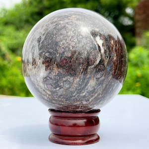 Puede incluir: Una piedra esférica pulida con un patrón complejo de remolinos gris oscuro, negro y blanco con inclusiones minerales de color rojo intenso. La esfera descansa sobre un soporte de exhibición circular de madera color marrón rojizo.