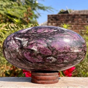Puede incluir: Una piedra grande, de forma ovalada, con una superficie pulida. La piedra presenta una mezcla de colores morado oscuro, negro y blanco, creando un efecto veteado. Descansa sobre un pequeño soporte redondo de madera. La piedra es un objeto decorativo.