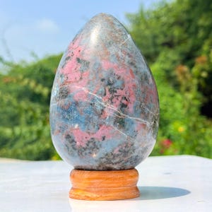 Op de afbeelding: Een gepolijst, eivormig stenen beeldhouwwerk met een gevlekt patroon van roze, grijze en blauwe mineraalinsluitingen. De steen is gemonteerd op een ronde, lichtbruine houten voet en heeft een glad, afgerond oppervlak met natuurlijke aders.