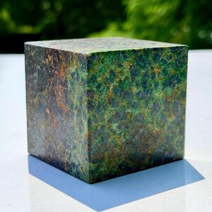 May include: A polished stone cube featuring a mottled texture with deep forest green, dark charcoal, and metallic bronze-colored mineral inclusions. The geometric object has sharp, clean edges and a smooth, reflective surface finish throughout.