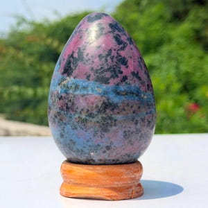 Op de afbeelding: Een gepolijst, eivormig stenen beeldhouwwerk met een gevlekt patroon van diep magenta, donkerzwart en levendige blauwe mineraalinklusionen. Het gladde, ronde object is bevestigd op een ronde, lichtbruine houten displaystandaard.