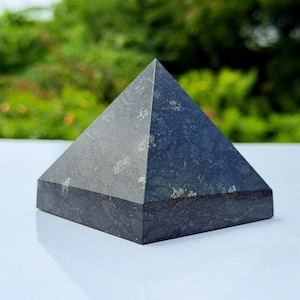 Op de afbeelding: Een donkergrijze stenen piramide met een gladde, gepolijste afwerking en zichtbare natuurlijke mineraalinsluitingen. Het object heeft een vierkante basis en vier driehoekige zijden die samenkomen in een scherpe top met een gevlekte textuur.
