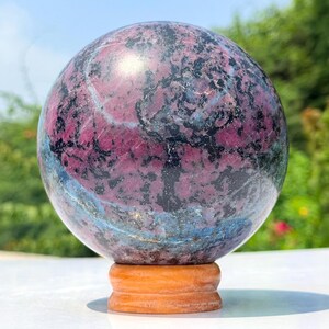 May include: A polished spherical stone featuring a mottled pattern of deep magenta, black, and hints of blue-grey mineral inclusions. The sphere rests securely on a small, circular, light-coloured wooden stand, showcasing a smooth, reflective surface.