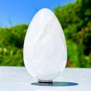 May include: A smooth, egg-shaped clear quartz crystal sculpture with a polished surface and internal natural inclusions. The crystal is mounted on a small, circular, dark metallic base that provides stability for the upright, vertical orientation.