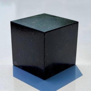 Könnte beinhalten: Ein massiver, dunkelschwarzer Würfel mit einer polierten, reflektierenden Oberfläche. Das Material weist eine subtile, natürliche Sprenkelung auf, die für eine texturierte Optik sorgt. Das geometrische Objekt hat scharfe Kanten und Ecken.
