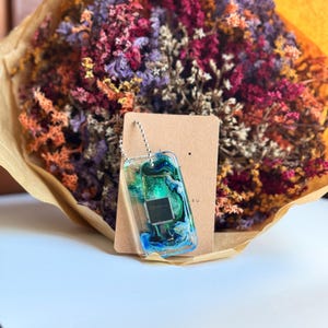 Może przedstawiać: Brelok z żywicy w kolorze niebieskim i zielonym z małym srebrnym chipem w środku. Brelok leży na brązowym papierowym tle z widocznym napisem "EU".