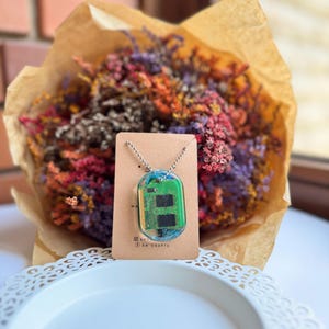 Może przedstawiać: Un unikalny naszyjnik z wisiorkiem z zieloną płytką drukowaną, zamkniętą w przezroczystej żywicy. Wisiorek wisi na srebrnym łańcuszku i jest prezentowany na brązowej karcie. Tło obejmuje bukiet kolorowych suszonych kwiatów.