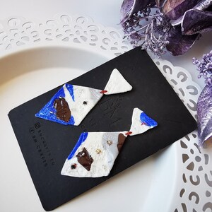 Könnte beinhalten: Ein Paar handgefertigte Ohrringe in geometrischer Diamantform. Die Ohrringe sind aus weißem Ton mit blauen, braunen und goldenen Akzenten. Die Ohrringe werden auf einem schwarzen Hintergrund mit dem Text "@BNCRAFTS_EU #BNCRAFTS" präsentiert.