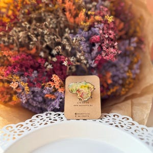 May include: A colorful pin in the shape of a country, with a floral design, is displayed on a brown card. The pin is set against a backdrop of dried flowers in shades of purple, orange, and pink.