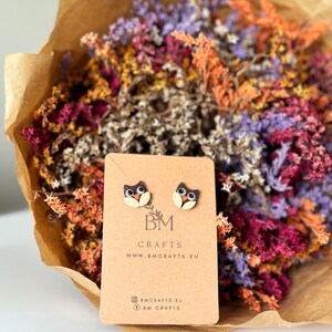 Könnte beinhalten: Ein Paar braune und weiße Eulen-Ohrstecker auf einer braunen Karte mit dem Text "BM CRAFTS" und "www.bmcrafts.eu". Die Karte enthält auch Social-Media-Symbole für Instagram und Facebook mit dem Text "@bmcrafts.eu" und "@bm crafts".