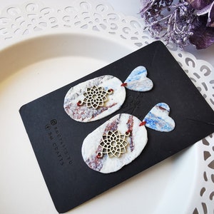 May include: A pair of handmade earrings with a white, blue, and red marbled design. Each earring features a metal filigree charm and a heart-shaped pendant.