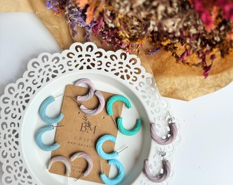 May include: A collection of colorful, handmade hoop earrings in shades of blue, pink, and green, displayed on a white, decorative plate. The earrings have a textured, twisted design. A brown tag with the text "BM CRAFT" is visible.