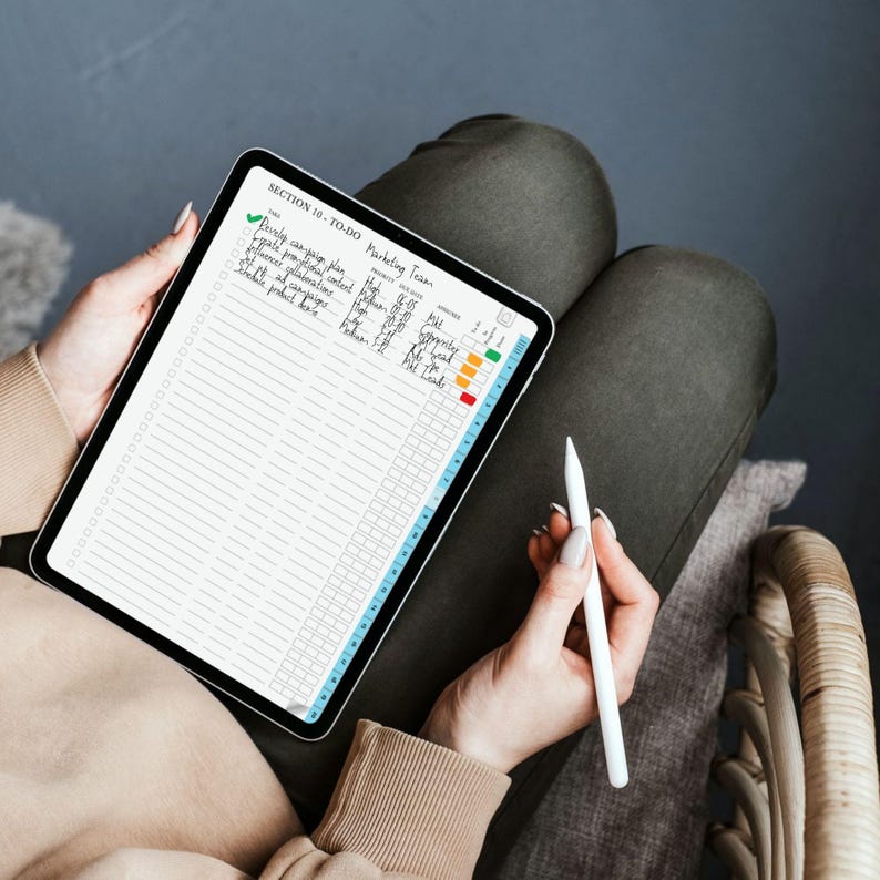 May include: A digital tablet displaying a to-do list with the text "SECTION 18 - TO-DO Marketing Team". The tablet is held by a person, with a stylus in hand. The screen shows a checklist with various tasks and color-coded progress indicators.