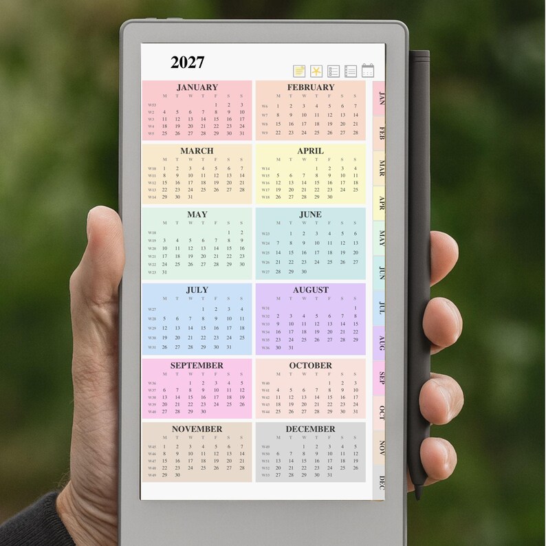 May include: A digital tablet displaying a 2027 annual calendar with twelve color-coded monthly grids. The device features a light gray frame and a black stylus attached to the side. The screen shows months from January to December.