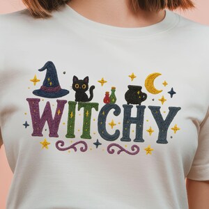 Peut inclure: T-shirt blanc avec le mot "WITCHY" brodé. Le motif comprend un chapeau de sorcière, un chat noir, des fioles de potion, un chaudron, des étoiles et un croissant de lune. Le design est dans les tons de bleu, vert, violet et or.