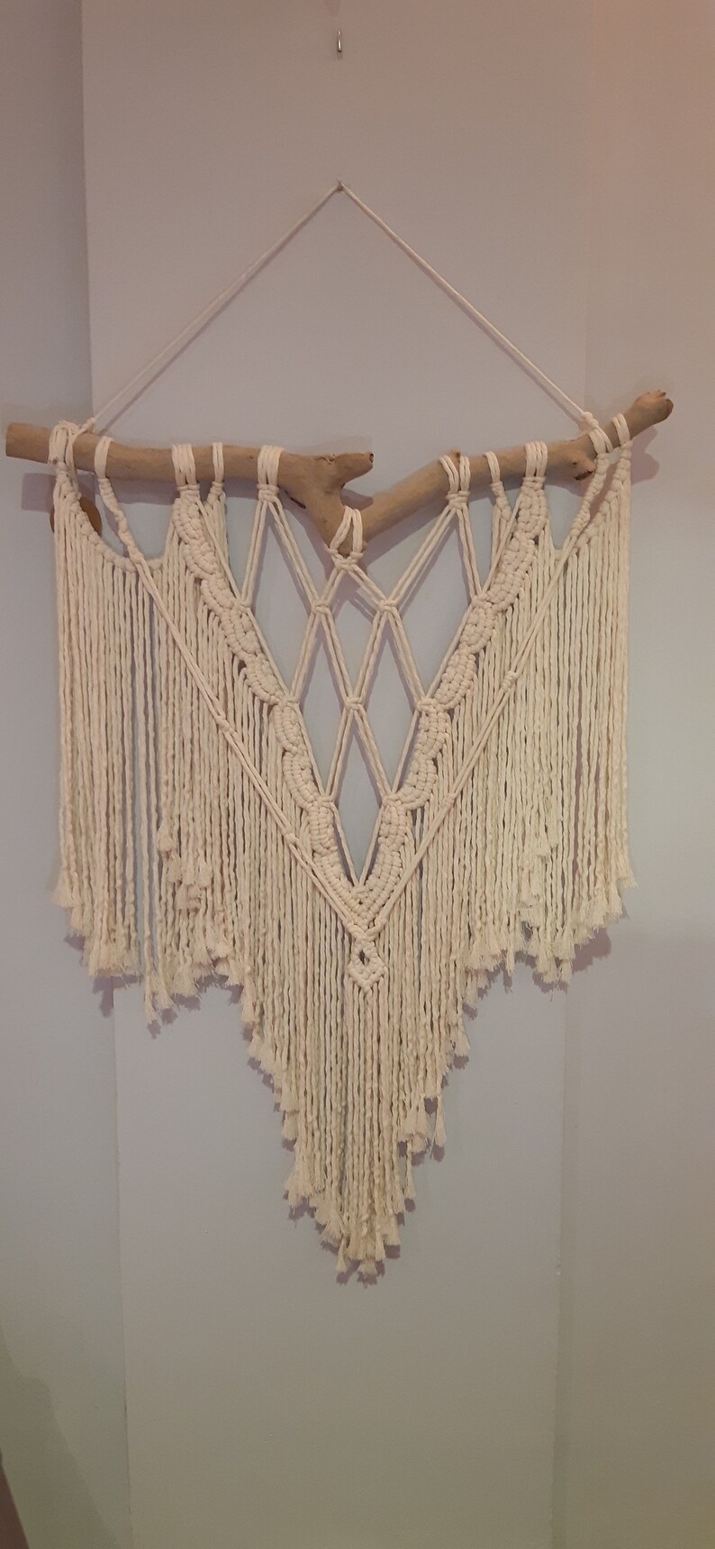 May include: A white macrame wall hanging with a driftwood branch at the top. The macrame is in a diamond shape with fringe hanging down the sides.