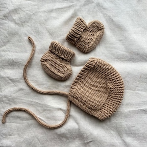 May include: A set of knitted baby accessories in a light brown color. The set includes a hat with a tie, and two booties.