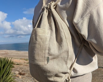 May include: A beige linen drawstring backpack with a small pocket on the front. The backpack is being worn by a person wearing beige pants.