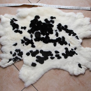 May include: A white and black spotted rabbit fur rug. The rug has a unique shape and a soft, fluffy texture. A measuring tape is placed across the top of the rug, indicating its size. The rug's pattern is reminiscent of a Dalmatian dog.
