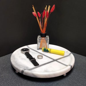 Op de afbeelding: Een wit marmeren dienblad met een grijs kruispatroon en een verhoogde rand. Het dienblad heeft drie zilveren voetjes en is versierd met een horloge, een gele lippenbalsem en een glazen fles met rode gedroogde bloemen.