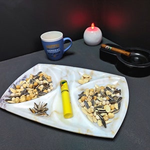 Puede incluir: Una bandeja de servir de mármol blanco con cuatro secciones, cada una llena de diferentes tipos de nueces y semillas. La bandeja está sobre una superficie negra con una taza azul que dice "Coffee Always Natural Fresh" y un cenicero negro con una vela encendida.