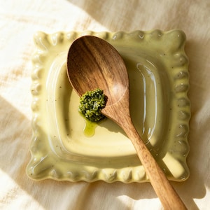 Könnte beinhalten: Ein Holzlöffel liegt auf einem quadratischen, hellgelben Keramikteller mit gewelltem Rand. Ein Klecks grünes Pesto liegt auf dem Teller, mit etwas Öl. Der Löffel ist braun und hat einen langen Griff.
