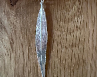 May include: A silver-toned pendant shaped like a long, slender leaf with a textured surface, suspended from a delicate matching silver-toned chain. The leaf features a prominent central vein and fine, realistic details across its entire surface.