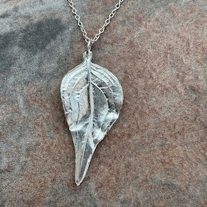 Puede incluir: Un collar de plata con un colgante en forma de hoja. El colgante está detallado con venas y una superficie texturizada.