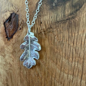Puede incluir: Un collar de tono plateado con un colgante en forma de hoja de roble con una superficie metálica texturizada. El colgante cuelga de una delicada cadena plateada con pequeños eslabones ovalados uniformes, mostrando un patrón de venas detallado.