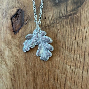 May include: A silver-colored necklace featuring a pendant shaped like two overlapping oak leaves with visible vein textures. The pendant hangs from a delicate silver-colored metal chain with small, uniform oval links, showcasing a metallic finish.