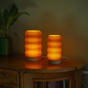 Pode incluir: Duas lâmpadas de papel laranja com um padrão ondulado em uma mesa de madeira. As lâmpadas estão acesas e projetam um brilho quente.