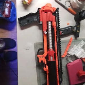 May include: A disassembled orange and black toy gun with a silver spring. The gun is lying on a white surface with other disassembled parts.
