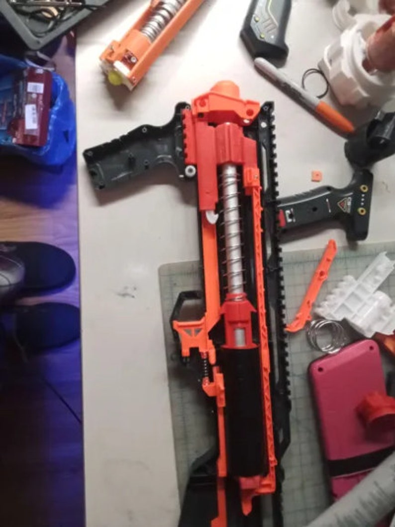 May include: A disassembled orange and black toy gun with a silver spring. The gun is lying on a white surface with other disassembled parts.