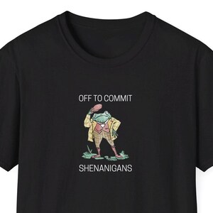 May include: Black t-shirt featuring the text "OFF TO COMMIT SHENANIGANS" and a cartoon frog in a tweed jacket, waistcoat, and boots, holding a hat. The frog is standing on green grass.