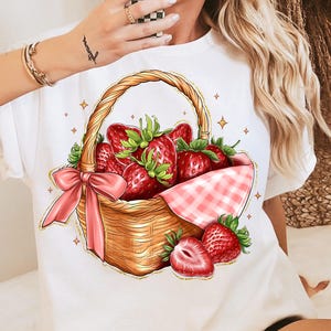 May include: White t-shirt with a graphic of a wicker basket filled with red strawberries and a pink gingham bow.