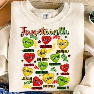 Puede incluir: Una sudadera blanca con la palabra "Juneteenth" en colores arcoíris. La sudadera presenta un diseño de corazones rojos, amarillos y verdes con texto dentro de cada corazón. El texto incluye nombres de afroamericanos notables y frases como "Dream", "Lead", "Fight", "Think", "Write", "Speak", "Dare", "Reclaim", "Inspire", "Win", "Challenge" y "Educate".