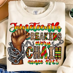 May include: A white sweatshirt with a colourful graphic design that says "Juneteenth Breaking Every Chain Since 1865". The graphic features a black fist breaking free from chains, a leopard print pattern, and colourful stripes.