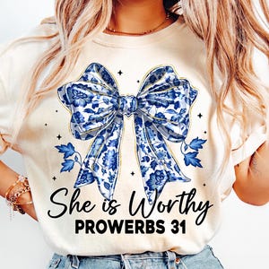 Op de afbeelding: Een wit T-shirt met een blauwe en gouden bloemen strik en de tekst "She is Worthy Proverbs 31".