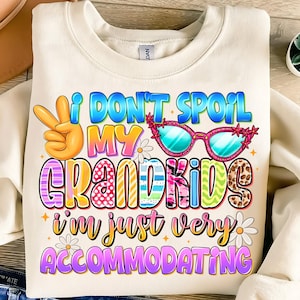 Op de afbeelding: Crèmekleurige sweatshirt met een kleurrijke afbeelding met de tekst "I DON'T SPOIL MY GRANDKIDS I'M JUST VERY ACCOMMODATING." Het ontwerp bevat een vredesteken, een zonnebril en madeliefjes. De tekst is in verschillende kleuren en lettertypen.