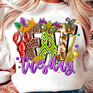 May include: White sweatshirt with a colorful graphic design. The design features the words "FAT Tuesday" in purple script, along with a lobster, crown, mask, fleur-de-lis, saxophone, and other Mardi Gras elements.