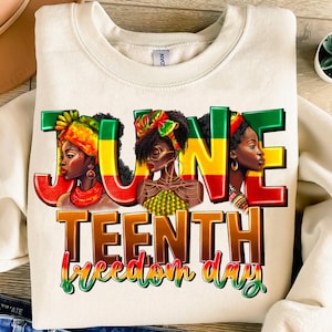 Puede incluir: Una sudadera blanca con un diseño gráfico que presenta las palabras "Juneteenth Freedom Day" en letras de colores. El diseño incluye tres mujeres negras con pañuelos de cabeza y joyas coloridas.
